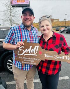 A smiling couple standing in a parking lot holding a wooden Sold sign Home Sweet Home, celebrating the successful sale of their home.